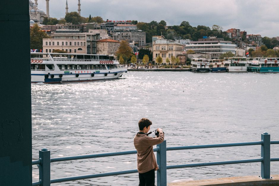 A young individual is taking photos by the Bosphorus in Istanbul, capturing local ferries and cityscape.